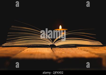 Close-up of the edge of the pages of an open book on an antique wooden plank table in semi-darkness with a lighted candle in the background. Stock Photo
