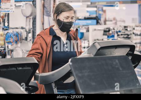 Serious woman in glasses and mask against virus buys large treadmill in a store Stock Photo