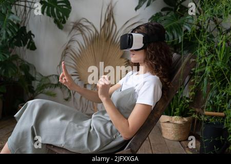 Young woman experiencing virtual reality while sitting among houseplants at greenhouse Stock Photo