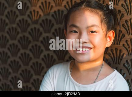 Portrait of cute Asian child girl with beautiful brown wooden wall, smiling face, natural sunlight, close-up image of healthy child girl, blank space Stock Photo