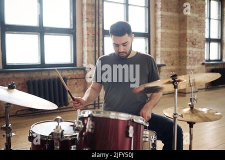 Young bearded man sitting at drum kit and performing on drums at studio during rehearsal Stock Photo