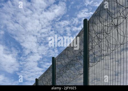 Metal security fence with razor wire on the top against a blue sky with white clouds. Stock Photo