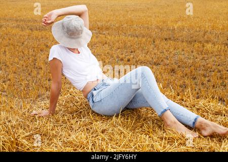 barefoot girl with straw hat sitting on a haystack on a bale in the agricultural field after harvesting. Stock Photo