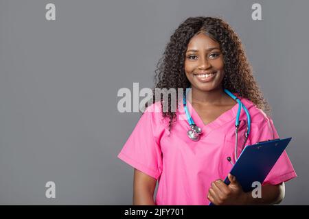 Young nurse using a clipboard against a grey background. Portrait Of Female Nurse with a stethoscope in pink uniform Stock Photo