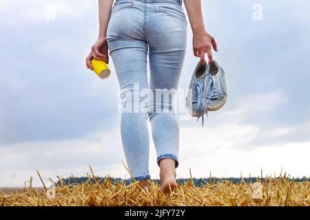 barefoot girl with sneakers and cardboard cup with coffee in hand standing in the agricultural field with haystack and bales after harvesting. Stock Photo