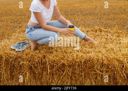 barefoot girl with sneakers and cardboard cup with coffee in hand standing in the agricultural field with haystack and bales after harvesting. Stock Photo