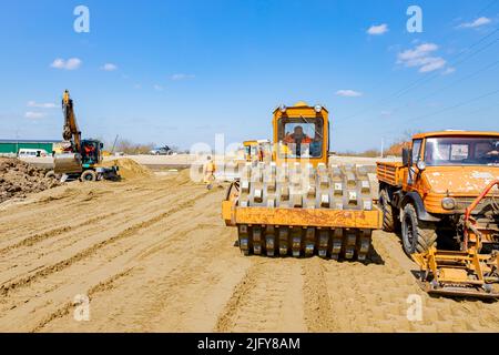 Road roller with spikes and truck with mounted plate vibration compactor are compacting, leveling sand for road foundation at building site. Stock Photo