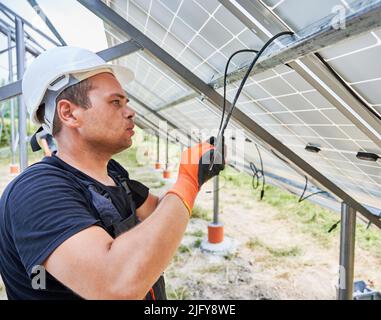 Worker fixing solar panels on metal beams with ropes. Electrician wearing helmet and workwear. Concept of renewable and ecological energy. Modern technology and innovation. Idea of environment safe Stock Photo