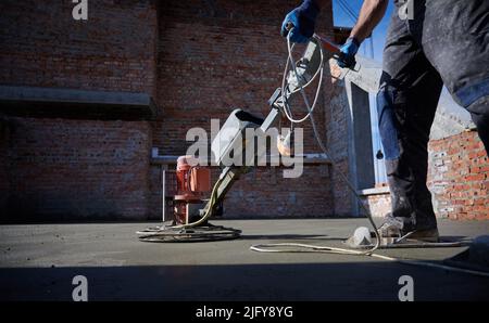 Close up male builder hands in protective gloves holding grout-grinding machine on concrete surface on the background of brick wall outside. Stock Photo