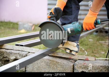 Worker cutting metal pole with grinder. Grinding machine. Partial image of man. Modern construction zone. Professional builder's uniform. Industrial gloves for hand protection in orange colour. Stock Photo