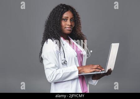 Portrait of a happy professional doctor at work. A young dark-skinned woman in a white laboratory coat with a stethoscope and a laptop stands in a med Stock Photo