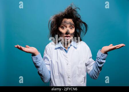 Mad scientist with funny expression and wacky hair, shrugging shoulders while on blue background. Silly looking crazy foolish chemist with dirty face lifting shoulders with ignorance. Studio shot. Stock Photo