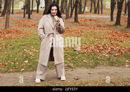 woman in a gray coat walking in the autumn park with a cup of hot drink. Stock Photo