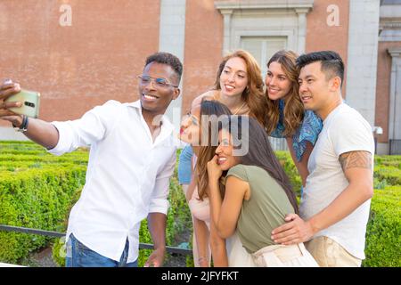 Multiracial group of friends taking a selfie with mobile int he street next to a garden Stock Photo