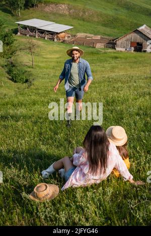 happy farmer in straw hat walking towards family sitting in picturesque meadow Stock Photo