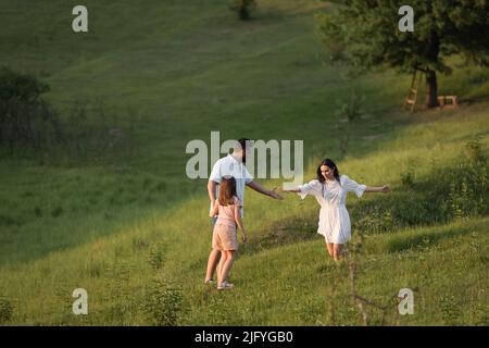 woman extending hand while running on grassy slope towards family Stock Photo