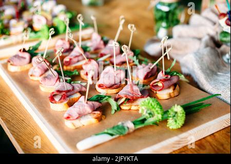 Delicious catering banquet buffet table decorated in rustic style in the garden. Different snacks, sandwiches with ham and greenery on a wooden plate. Stock Photo