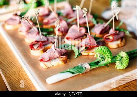 Delicious catering banquet buffet table decorated in rustic style in the garden. Different snacks, sandwiches with ham and greenery on a wooden plate. Stock Photo