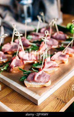 Delicious catering banquet buffet table decorated in rustic style in the garden. Different snacks, sandwiches with ham and greenery on a wooden plate. Stock Photo