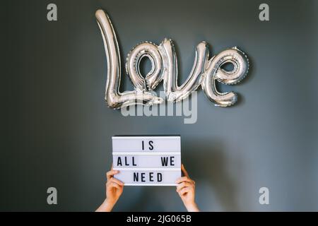 Male hands holding lightbox with Text Love is all we need under silver foil balloon on gray wall background. Film effect toning. Valentine's day conce Stock Photo