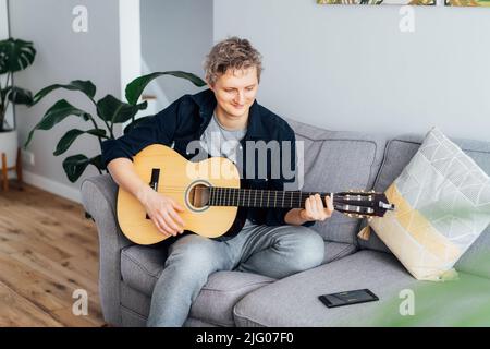 Man taking online guitar lessons via a mobile application on his phone at home sitting on the gray couch, sofa in the living room. He watching acousti Stock Photo