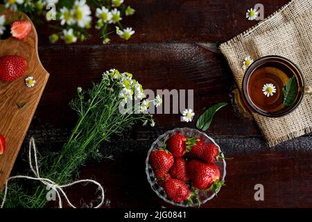 Fresh strawberries, summer chamomile flowers, herbal tea and mint on wooden table. Atmospheric feminine summer lifestyle. Beautiful food. Flat lay food. Stock Photo