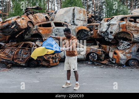 African man with Ukrainian flag stands against cars cemetery in Irpin near Kyiv out of many burnt and shelled cars. Consequences of the Russian milita Stock Photo