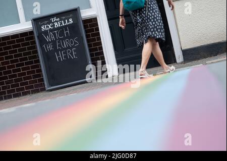 London, UK. 06th July, 2022. Soccer: National team, women, European Championship 2022: A woman walks past a sign that reads 'Watch the Euros here', which is placed in front of a pub. In the foreground, a rainbow is painted on the street. Credit: Sebastian Gollnow/dpa/Alamy Live News Stock Photo