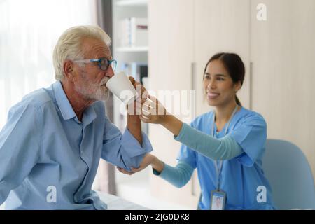Asian elderly caregiver woman sitting on a hospital bed next to an older man helping drink a glass of water from the bedroom at home. Stock Photo