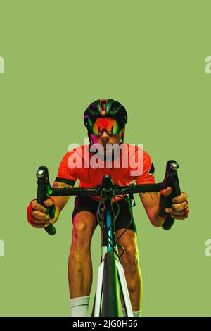 Front camera view of sportsman, cyclist on bicycle in red sports uniform and protective helmet isolated on green background. Stock Photo