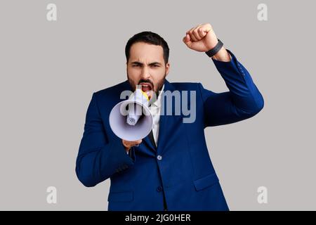 Portrait of bearded businessman standing with raised hands and holding megaphone, screaming in loud speaker, protesting, wearing official style suit. Indoor studio shot isolated on gray background. Stock Photo