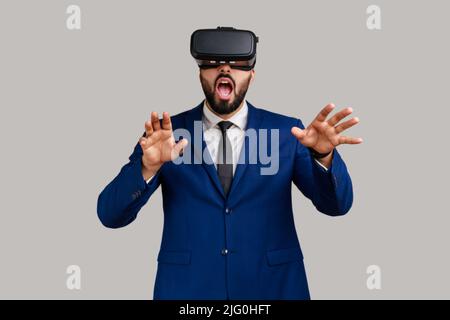 Bearded man in vr headset, playing virtual reality game with shocked facial expression, outstretching hands, wearing official style suit. Indoor studio shot isolated on gray background. Stock Photo