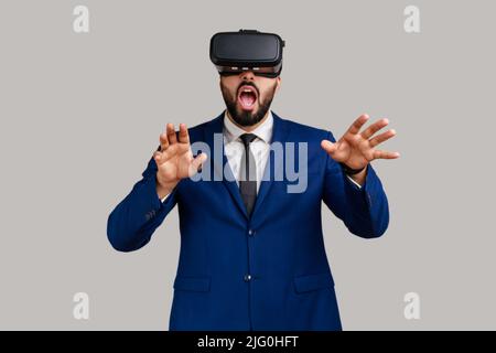 Bearded man in vr headset, playing virtual reality game with shocked facial expression, outstretching hands, wearing official style suit. Indoor studio shot isolated on gray background. Stock Photo