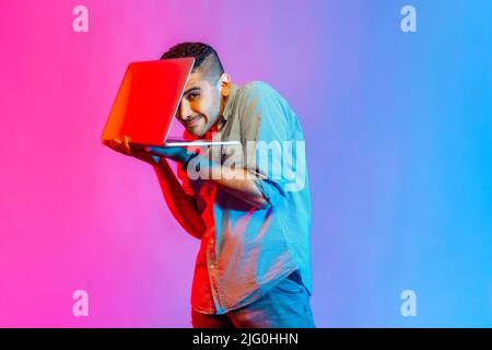 Portrait of funny young adult man in shirt holding laptop and looking at camera, spying, hiding his face behind notebook. Indoor studio shot isolated on colorful neon light background. Stock Photo