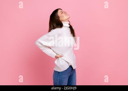 Unhealthy sick female touching back, suffering pain in lower lumbar muscular, kidney inflammation, wearing white casual style sweater. Indoor studio shot isolated on pink background. Stock Photo