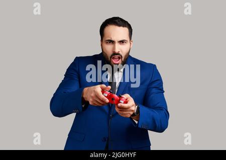 Concentrated attractive bearded man holding in hands red gamepad joystick, grimacing playing video games, wearing official style suit. Indoor studio shot isolated on gray background. Stock Photo