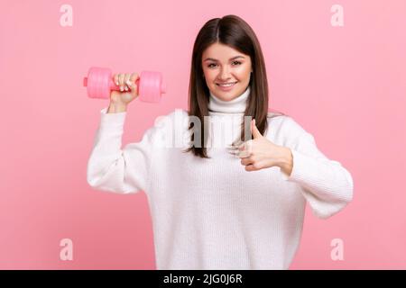 Positive smiling female holds rosy dumbbells in hand, showing thumb up, recommend to go in for sport, wearing white casual style sweater. Indoor studio shot isolated on pink background. Stock Photo