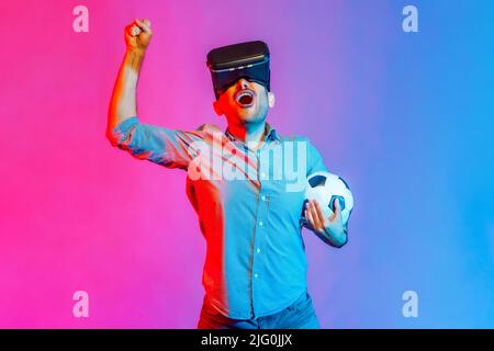 Portrait of man in shirt wearing headset and holding soccer ball, watching 3d football team, cheering, celebrating victory. Indoor studio shot isolated on colorful neon light background. Stock Photo