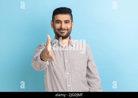 Portrait of handsome happy businessman standing and looking at camera with toothy smile and giving hand to greeting or handshake, wearing striped shirt. Indoor studio shot isolated on blue background. Stock Photo