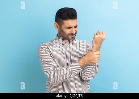 Unhappy sick bearded businessman touching painful hand, suffering trauma, sprain wrist, feeling ache of carpal tunnel syndrome, wearing striped shirt. Indoor studio shot isolated on blue background. Stock Photo