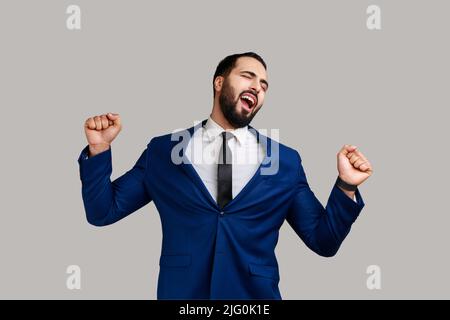 Portrait of sleepy tired exhausted bearded businessman standing and yawning with closed eyes and raised arms, wearing official style suit. Indoor studio shot isolated on gray background. Stock Photo