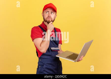 Portrait of bearded pensive worker man standing and working on laptop, taking orders, online support, holding chin, thinking about solution. Indoor studio shot isolated on yellow background. Stock Photo
