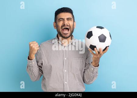 Bearded businessman screaming widely opening mouth, celebrating victory of favourite football team, holding soccer ball in hands, wearing striped shirt. Indoor studio shot isolated on blue background. Stock Photo