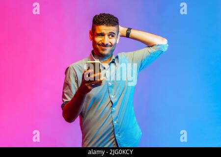 Portrait of man in shirt scratching head holding smartphone in hand, contemplating about software updating, choosing suitable tariffs. Indoor studio shot isolated on colorful neon light background. Stock Photo