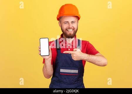 Satisfied builder man or repairman in uniform and protective helmet showing cell phone with blank empty screen with copy space, advertise his service. Indoor studio shot isolated on yellow background. Stock Photo