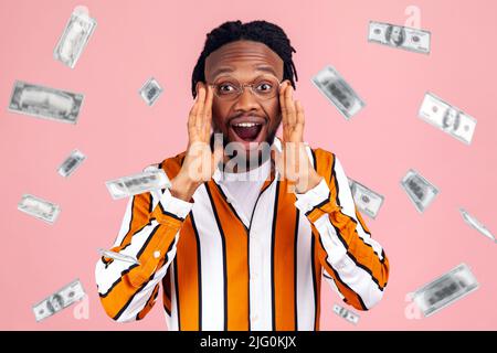 Money rain wow. Bemused surprised rich man looking at camera with overwhelming big eyes, shocked with amazing face. dollars money falling. Indoor studio shot isolated on pink background. Stock Photo