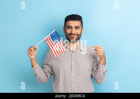 Portrait of happy positive man with beard standing and pointing at american flag, celebrating national holiday, wearing striped shirt. Indoor studio shot isolated on blue background. Stock Photo
