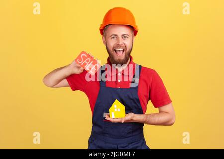 Portrait of happy smiling builder man holding paper house and card with sale inscription, offering to buy new apartment at low price. Indoor studio shot isolated on yellow background. Stock Photo