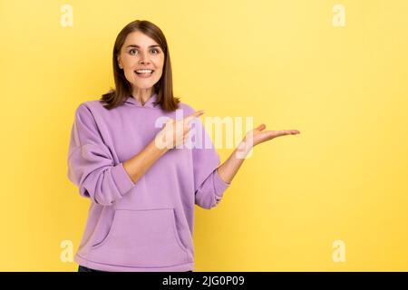 Portrait of cheerful young woman pointing at copy space on her palm, empty place for idea presentation, product advertising, wearing purple hoodie. Indoor studio shot isolated on yellow background. Stock Photo