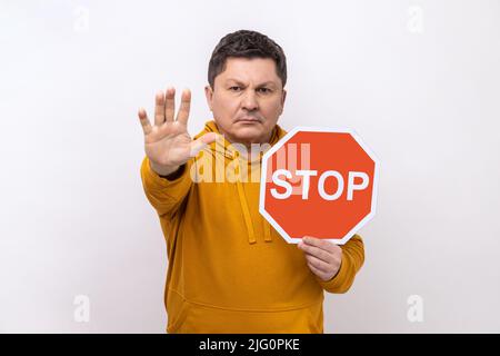 Concentrated serious man looking angrily showing stop gesture, holding road traffic sign, warning of ban, forbidden access, wearing urban style hoodie. Indoor studio shot isolated on white background. Stock Photo